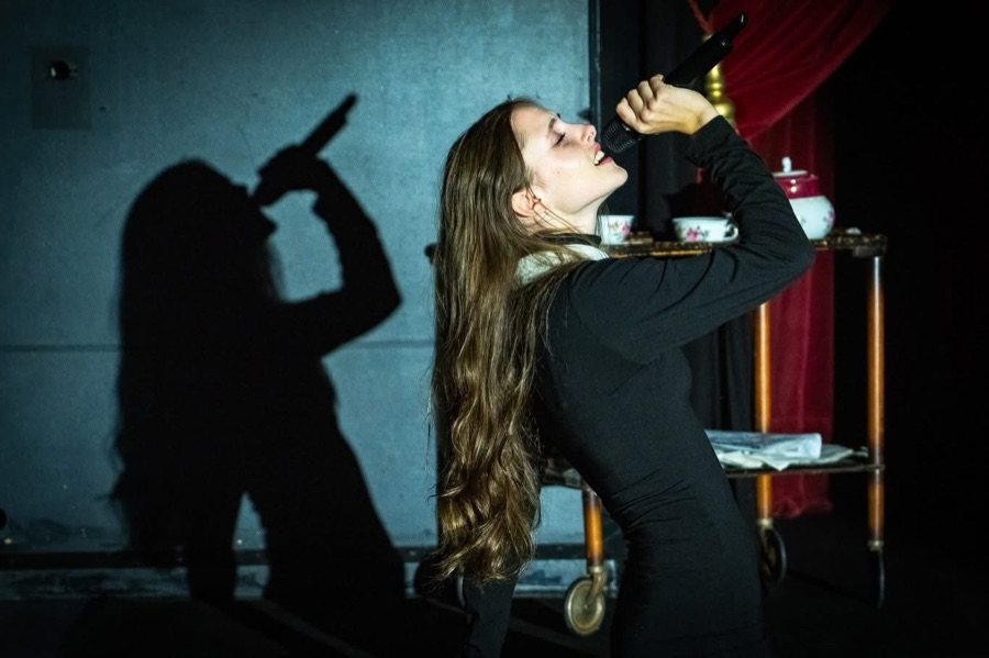 Susanne sings into a microphone, her shadow thrown large on the wall beside a tea trolley.