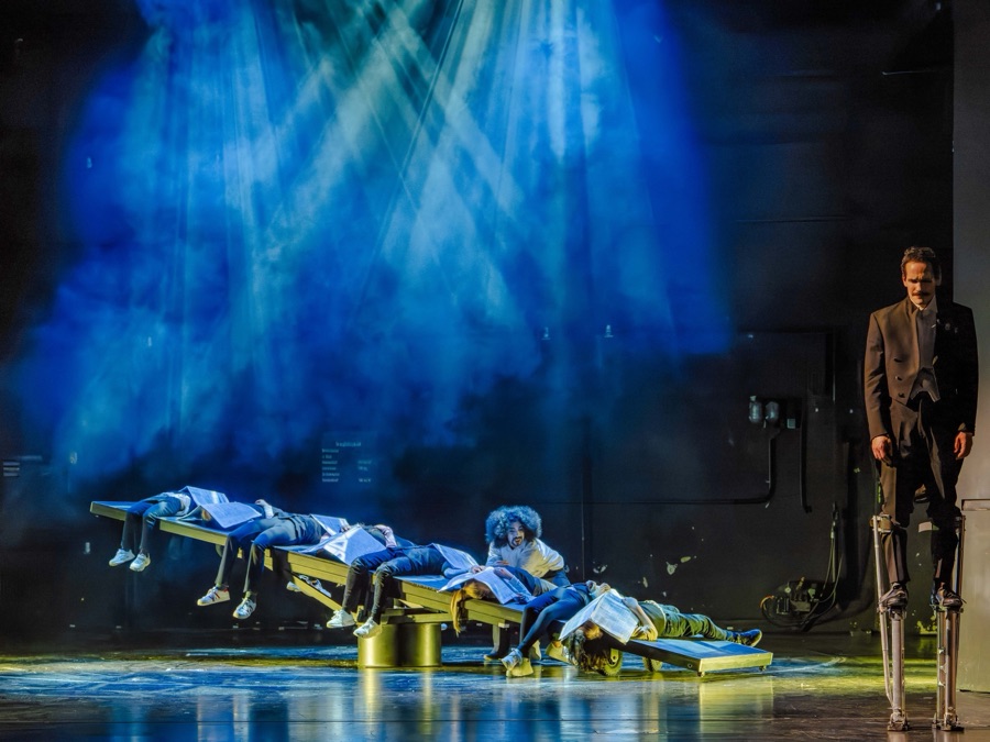 Children lie across a yellow seesaw under crossing beams of blue light while the clown watches on stilts.