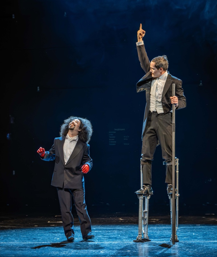 The clown master on stilts points to the sky while his wild-haired pupil looks on.