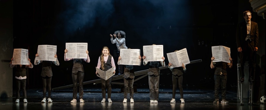 Children in a line hold up newspapers across the wide stage.