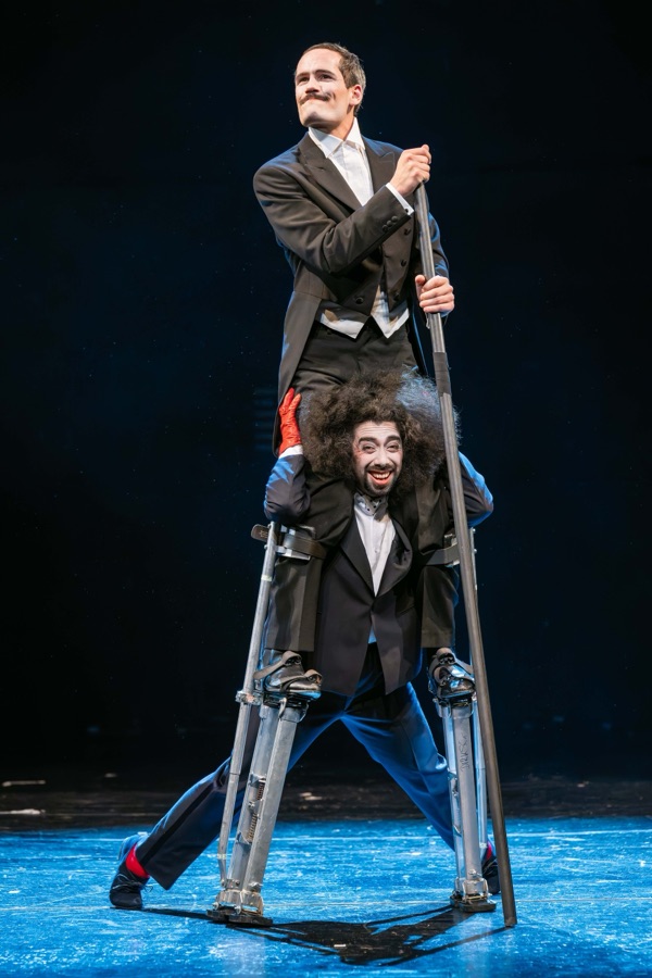 The clown in tailcoat towers on stilts above his pupil.