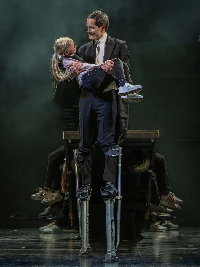 The clown on stilts carries a child in his arms above a bench.
