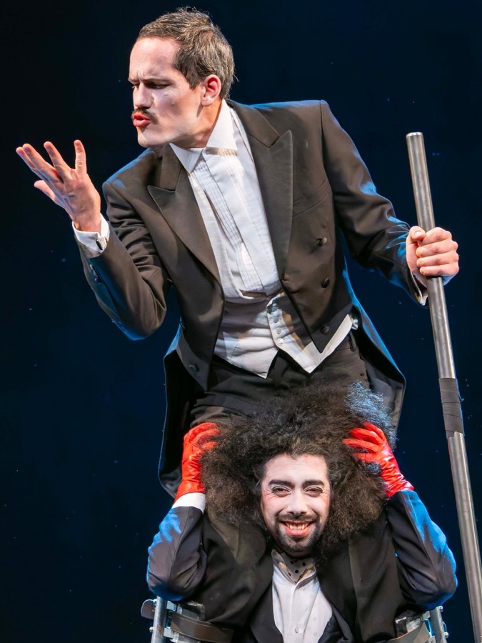 Close-up of the clown master in tailcoat, white face and painted moustache, gesturing theatrically while gripping the curly hair of the grinning dancer in red gloves below him.