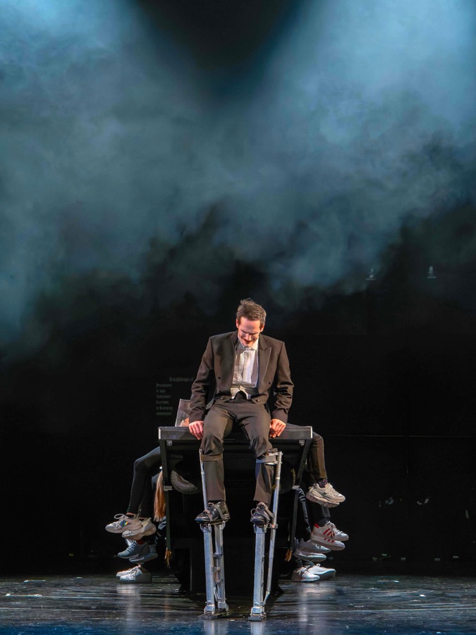The clown sits slumped on a bench on stilts under a cloud of stage fog, with children's legs protruding from under the bench like a many-legged creature.