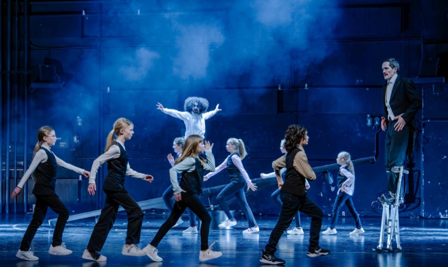 Children march in a line across the blue-lit foggy stage, the dancer in white poses with outstretched arms behind them and the clown stands on stilts at the right.