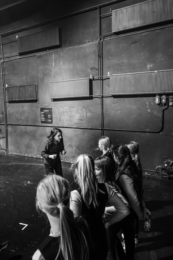 Black and white rehearsal photo: Petra Rațiu, fists clenched mid-explanation, works with the children of the Theaterkidsclub on the bare stage.