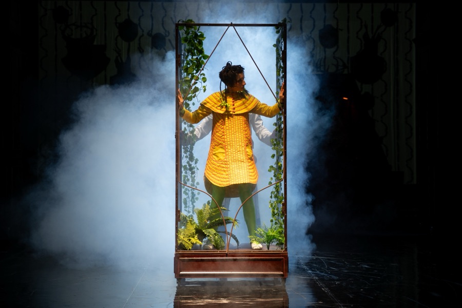Heidi stands inside a tall glass cabinet overgrown with ivy, surrounded by fog.