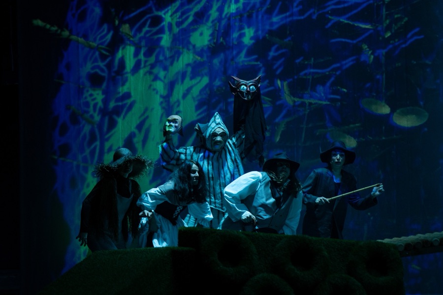 Masked figures and an owl gather on the mountain set in blue and green light.