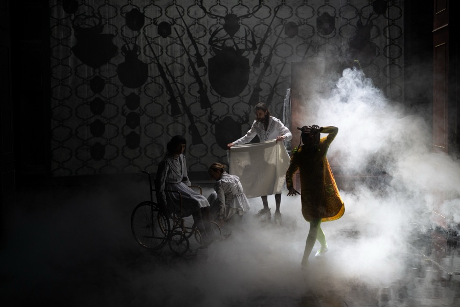 Figures moving through thick white stage fog in front of a wall of dark hunting trophies.