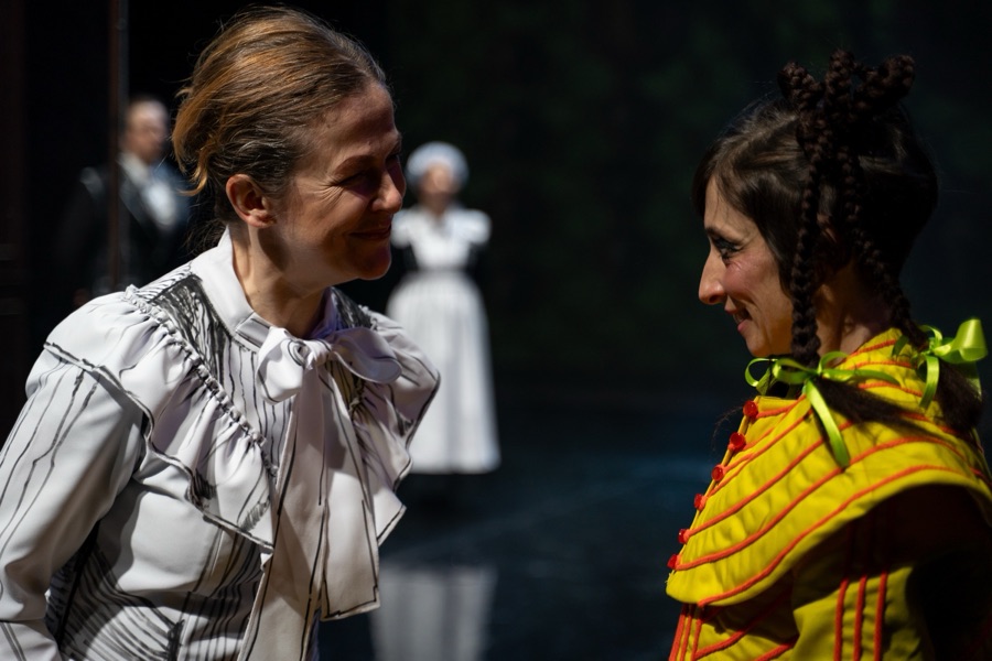 Miss Rottenmeier and Heidi, in her yellow quilted dress with green ribbons, face each other smiling.