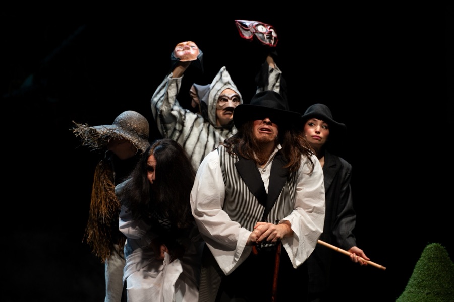 Masked villagers huddle together, holding masks on sticks above their heads.