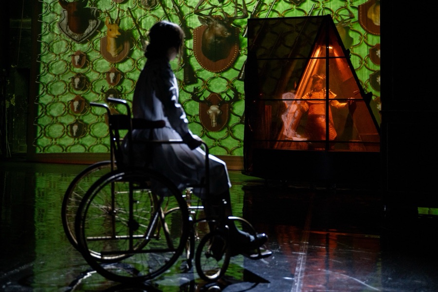 Klara in her wheelchair, seen from behind, before a wall of green projected wallpaper and a glowing doorway.