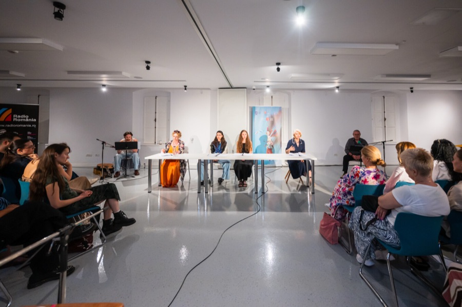 Wide view of the reading performance at Casa Artelor in Sibiu: actors seated behind a long white table with microphones in a bright white room, a further reader seated at a music stand and a Radio Romania banner at the left, and the audience seated on both sides in the foreground.