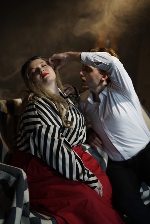 He leans over her and touches her forehead as she sits with her eyes closed, smoke rising behind them.