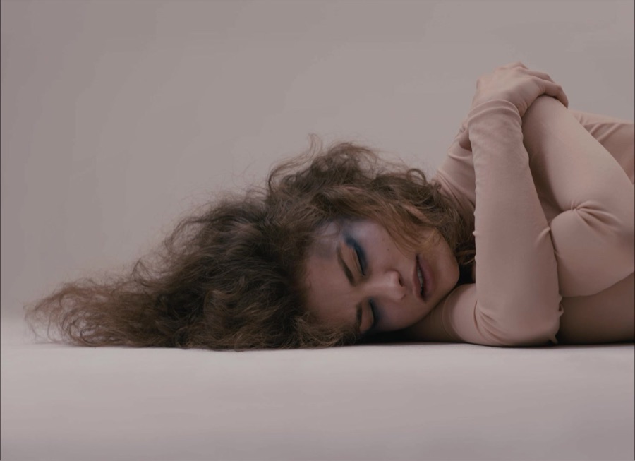 The dancer lies curled on her side on a pale studio floor, hair spread around her head.