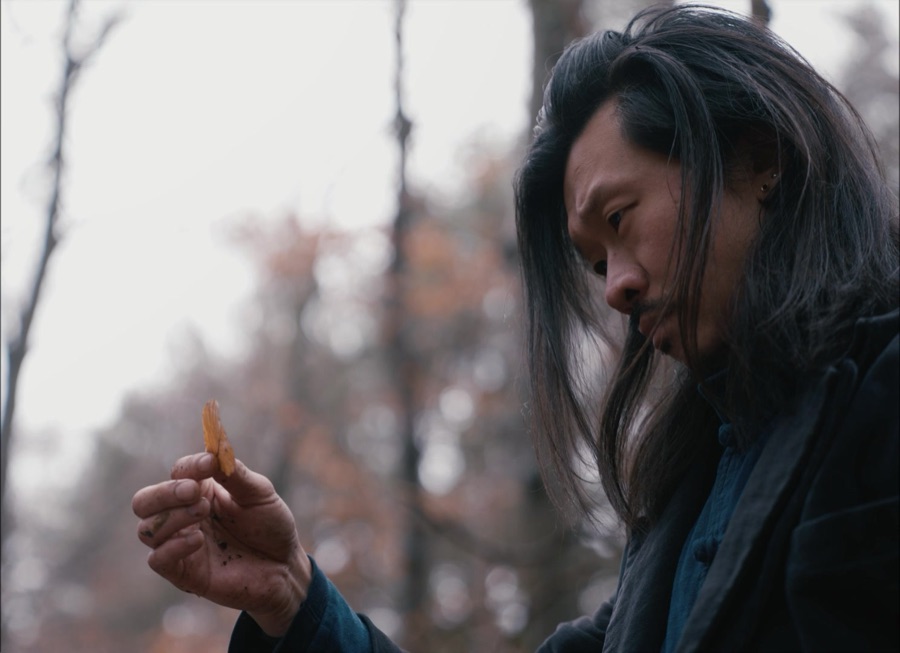 He studies a dry leaf held between his fingers, bare trees behind him.