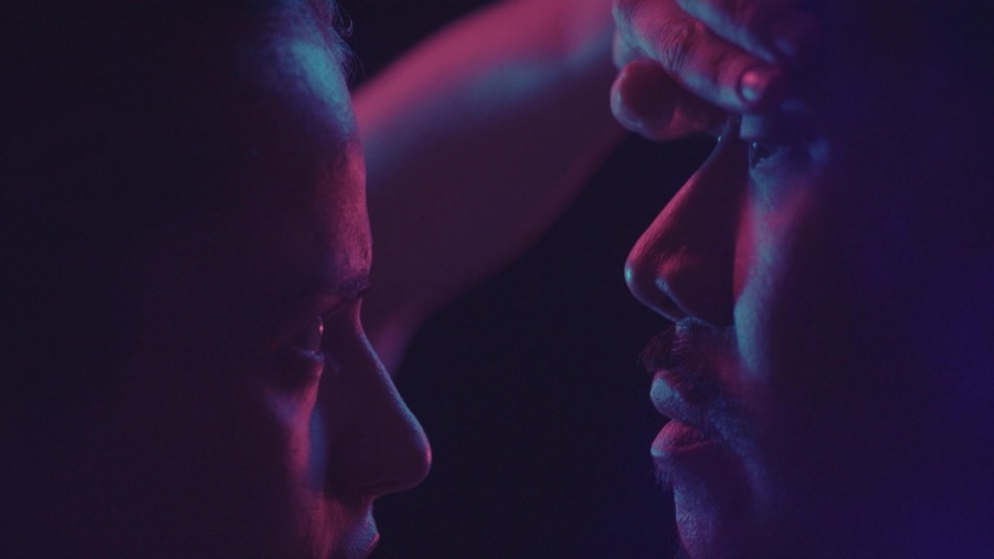 A hand rests on a forehead, faces in profile in deep violet light.
