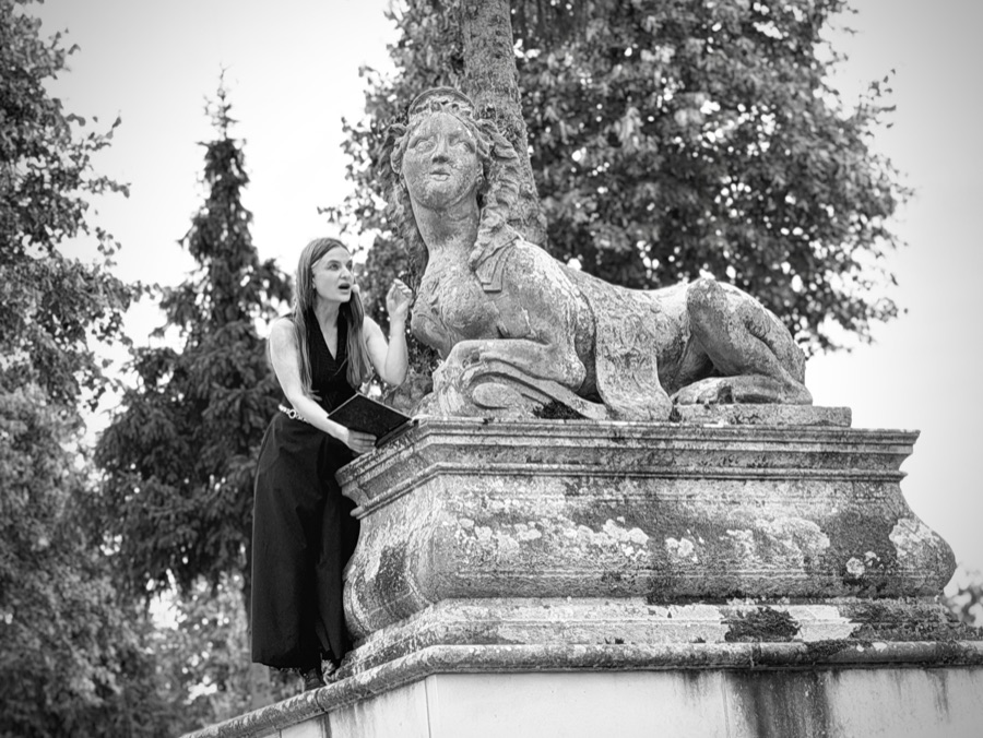 Black-and-white photo of AnniKa von Trier reading from a book while looking up at a stone sphinx on its pedestal.