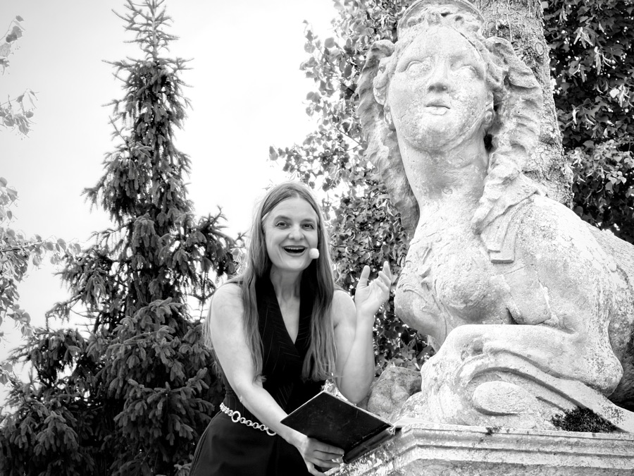 Black-and-white close-up of AnniKa von Trier laughing with an open book next to the weathered stone sphinx.