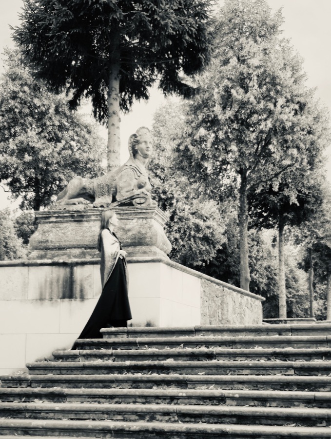 Black-and-white photo of AnniKa von Trier in a long dark dress standing beside a stone sphinx on the Sphinx staircase in the Rheinsberg palace garden.