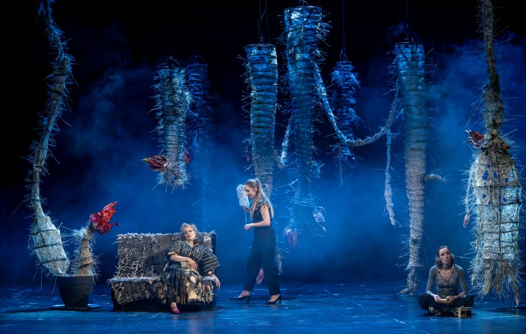 Wide view of Cristina Milea's set: giant thorny cactus-like forms hang in blue fog while Lin sits on the sofa, Clara stands beside her and Rahel sits cross-legged on the floor.