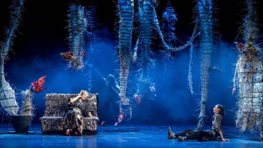 The set in blue haze: thorny sculptures hang from above, Lin on the sofa, Rahel lying on the floor.