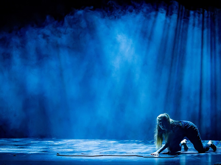 A woman crawls across the bare stage under a wash of blue light.