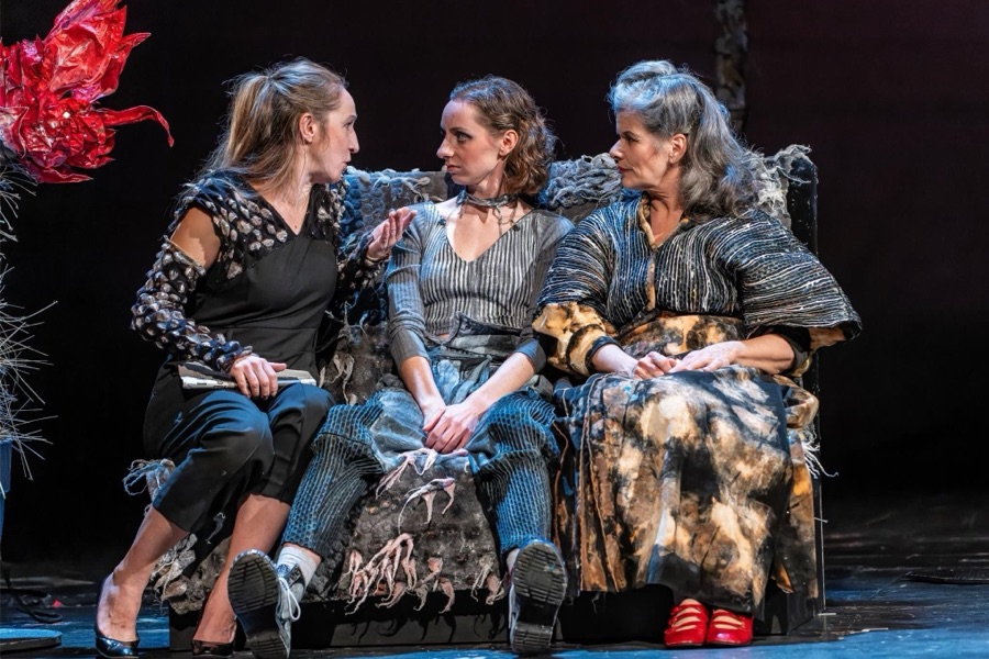 Clara, Rahel and Lin sit close together on the shaggy sofa looking at one another, a red paper flower on a spiky cactus sculpture at the left.