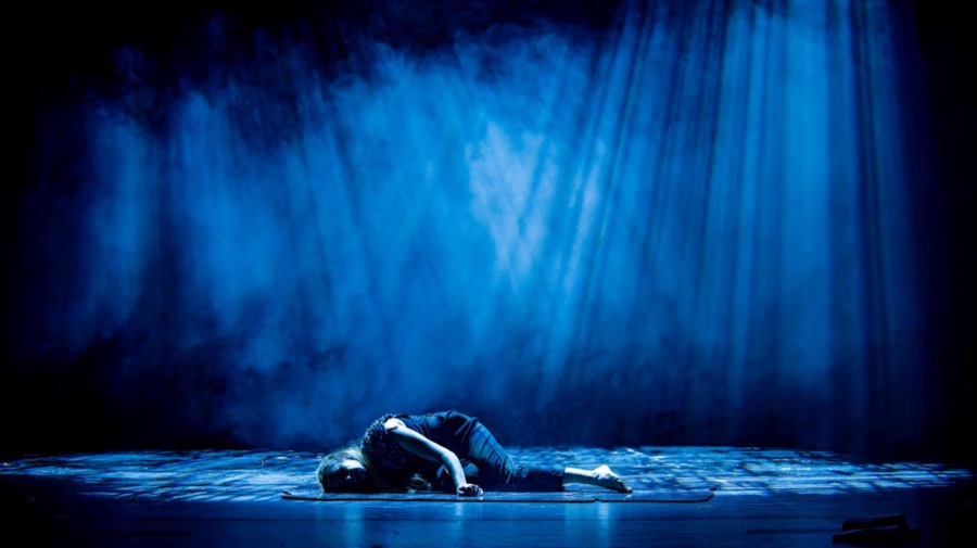 A lone figure in black lies curled on the bare stage floor under a wash of blue light and fog.