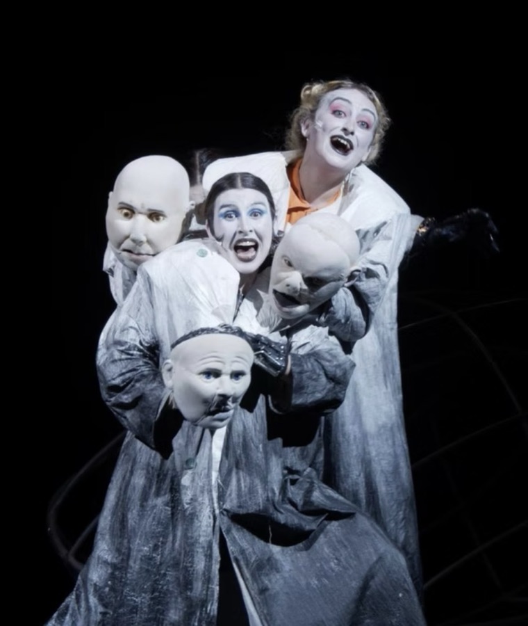 Three performers with white-painted faces laugh from inside a cluster of bald puppet heads and grey coats.