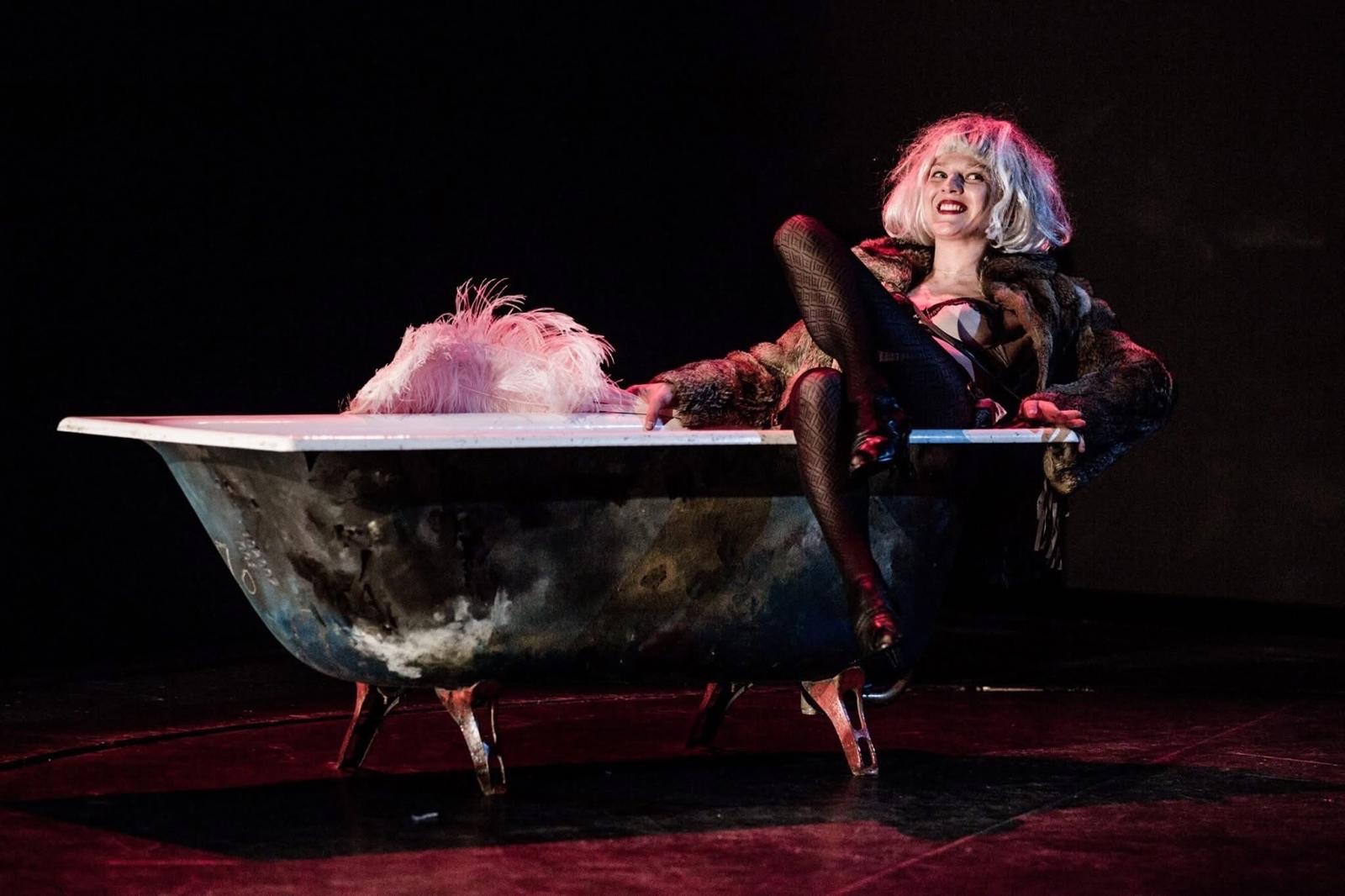 A woman in a blonde wig and fur reclines in a bathtub, one leg over the rim, smiling at the audience.