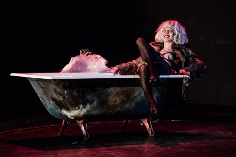 A woman in a blonde wig and fur reclines in a bathtub, one leg over the rim, smiling at the audience.