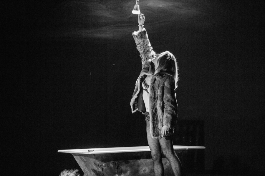 Black and white: a figure in a fur coat stands in the bathtub reaching up to a hanging lamp.