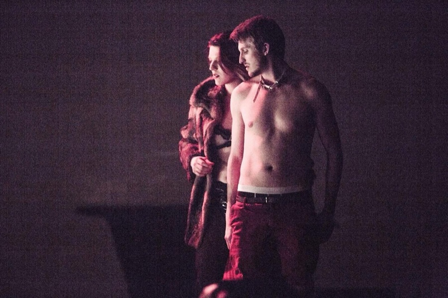 The woman in fur stands behind the bare-chested man, who wears a collar, in red light.